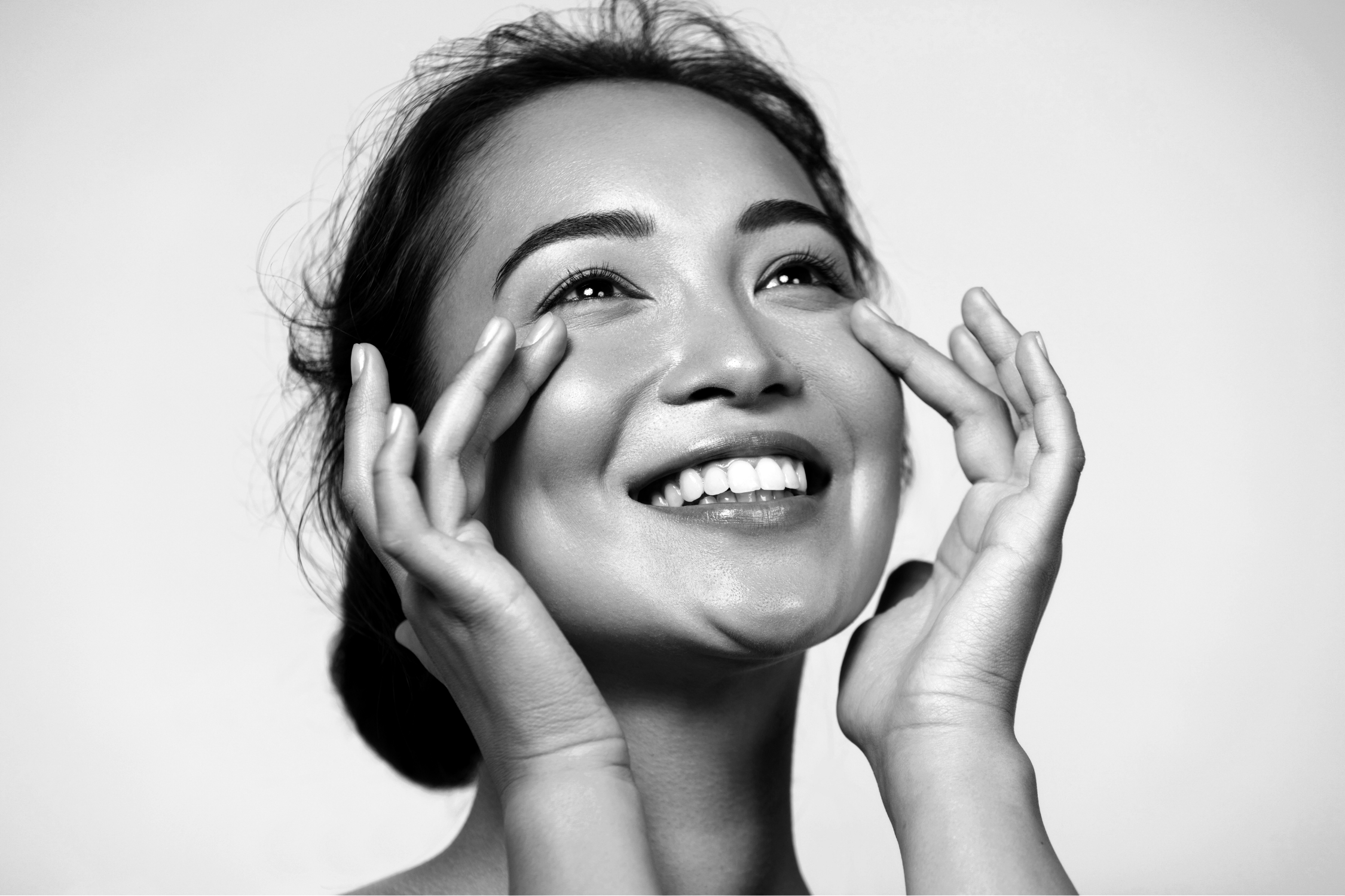 Black and white image of a young woman smiling while applying skincare to heer lower eyelids