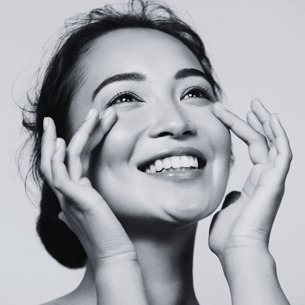 Woman with clear skin smiling joyfully, raising hands to face, monochrome background. Emphasis on her bright expression and radiant eyes.