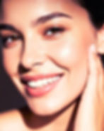 Close-up of a smiling person with glowing skin, brown eyes, and dark hair. Hand touching cheek. Bright and warm lighting.