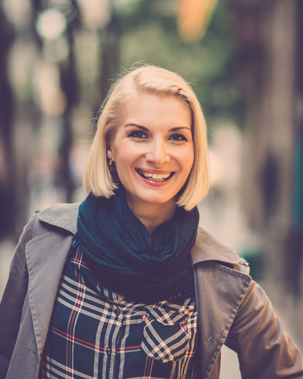 Smiling blonde woman in a plaid shirt, dark scarf, and coat stands outdoors against a blurred street and trees.
