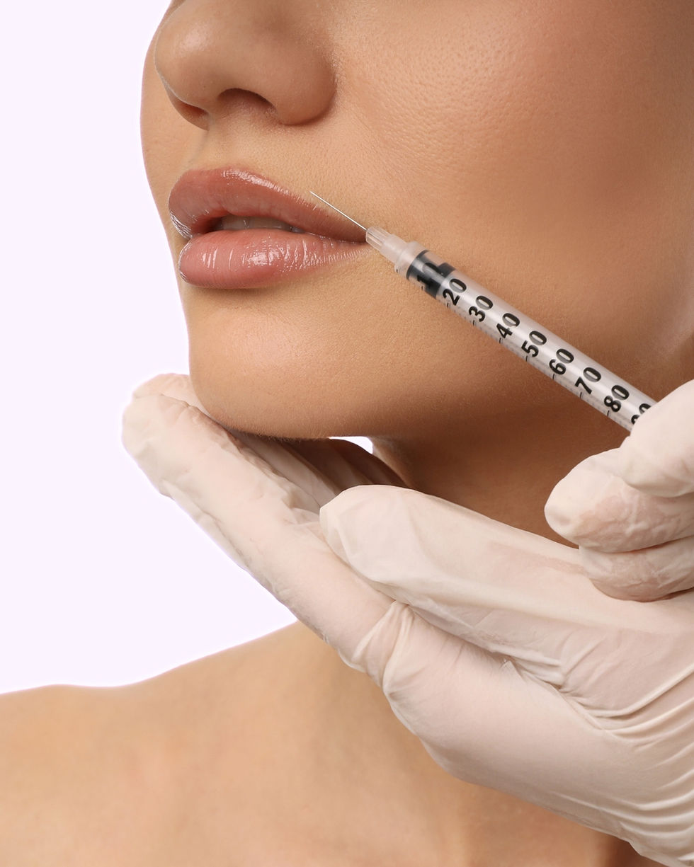 Close-up of a syringe near a woman's lips being held by gloved hands, suggesting a cosmetic procedure. The background is pale.