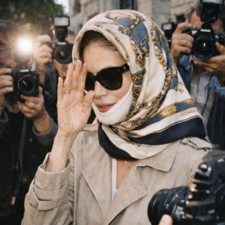 Woman in sunglasses and patterned headscarf shields face from paparazzi flashes. Background features stone building and photographers.