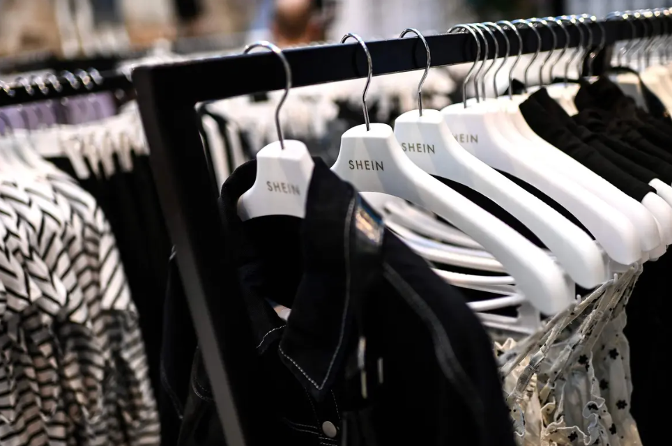 Clothes on Shein-branded white hangers are arranged on a rail in a store. Black and white garments with patterns are visible.