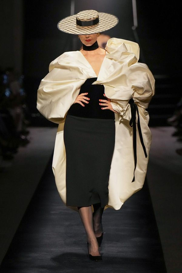 Model on runway in dramatic monochrome outfit with wide-brimmed hat, voluminous cream coat, and sleek black dress; elegant setting.