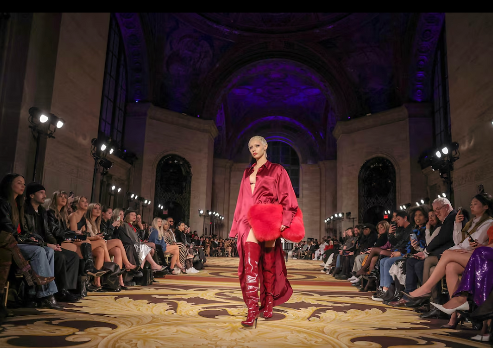 Model in red dress and boots walks runway in grand hall with arched ceiling. Audience surrounds, vibrant lighting creates elegant mood.