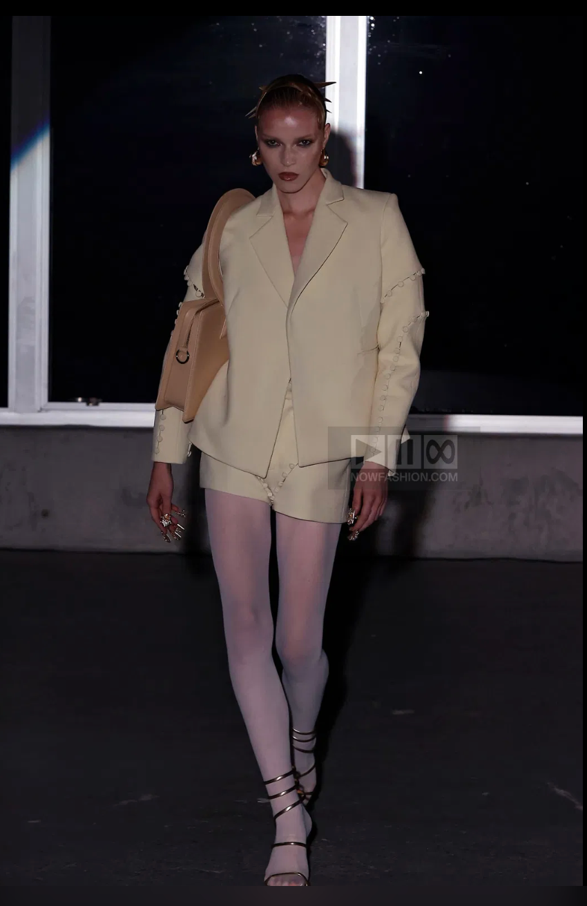 Model in pale yellow suit and shorts, white tights, strappy heels, holding a tan bag. Dark background, runway setting. Elegant mood.