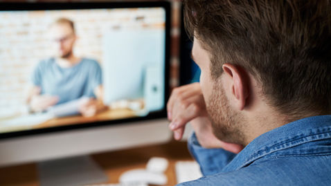 brown haired man at computer watching another man make something at a desk.