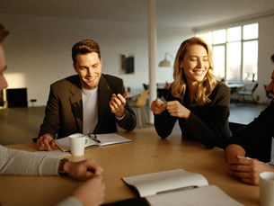Photographic. Three or four colleagues around a table in a bright office in warm late-morning light, leaning in, animated and engaged in the middle of a working conversation, notebooks and coffee, one person mid-sentence with hands moving. Documentary, warm grade, shallow depth of field, genuine energy, no screens dominating the frame.
