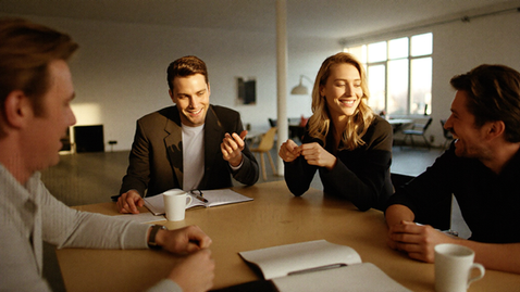 Photographic. Three or four colleagues around a table in a bright office in warm late-morning light, leaning in, animated and engaged in the middle of a working conversation, notebooks and coffee, one person mid-sentence with hands moving. Documentary, warm grade, shallow depth of field, genuine energy, no screens dominating the frame.