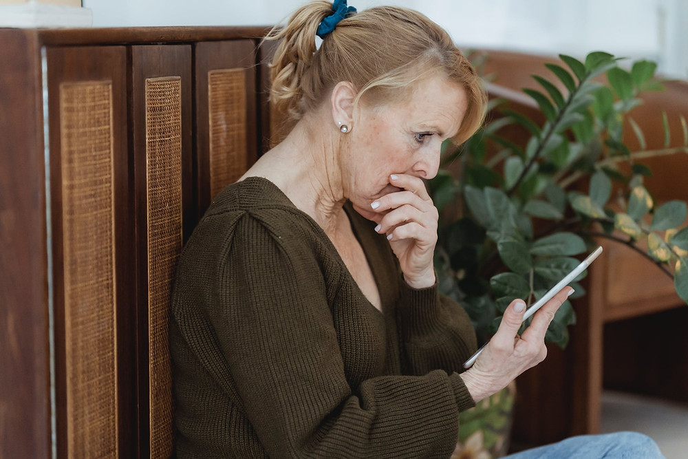 A middle aged woman sits on the floor of her living room with her back against a cabinet. She holds her hand to her mouth as she looks worriedly to her tablet that she holds with her other hand.