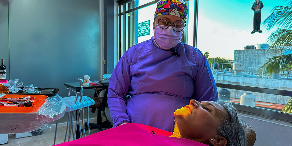 Dentista con bata morada y mascarilla atiende a una paciente reclinada con babero rosa en una clínica luminosa.