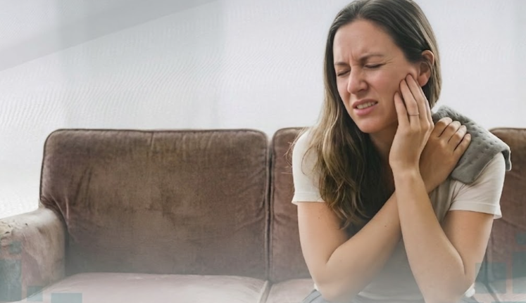 Mujer sentada en un sofá, con gesto de dolor y mano en la mejilla, en un salón luminoso.