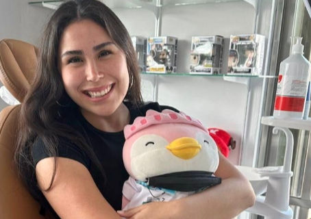 Mujer sonriente abraza un peluche de pingüino en una clínica dental, con estantes y gel desinfectante al fondo.