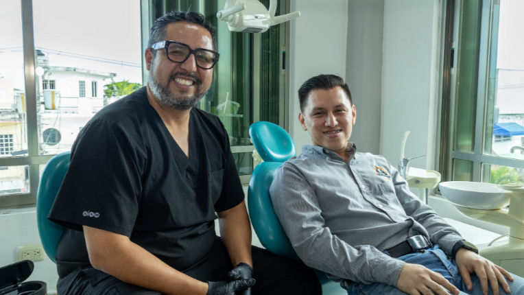 Dos hombres sonrientes en una clínica dental, uno con uniforme negro y otro recostado en silla azul junto a equipos dentales.