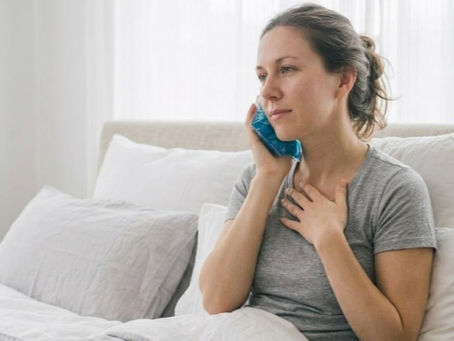 Mujer en reposo en la cama, con una compresa fría en la mejilla y la mano en el pecho, en un dormitorio blanco.