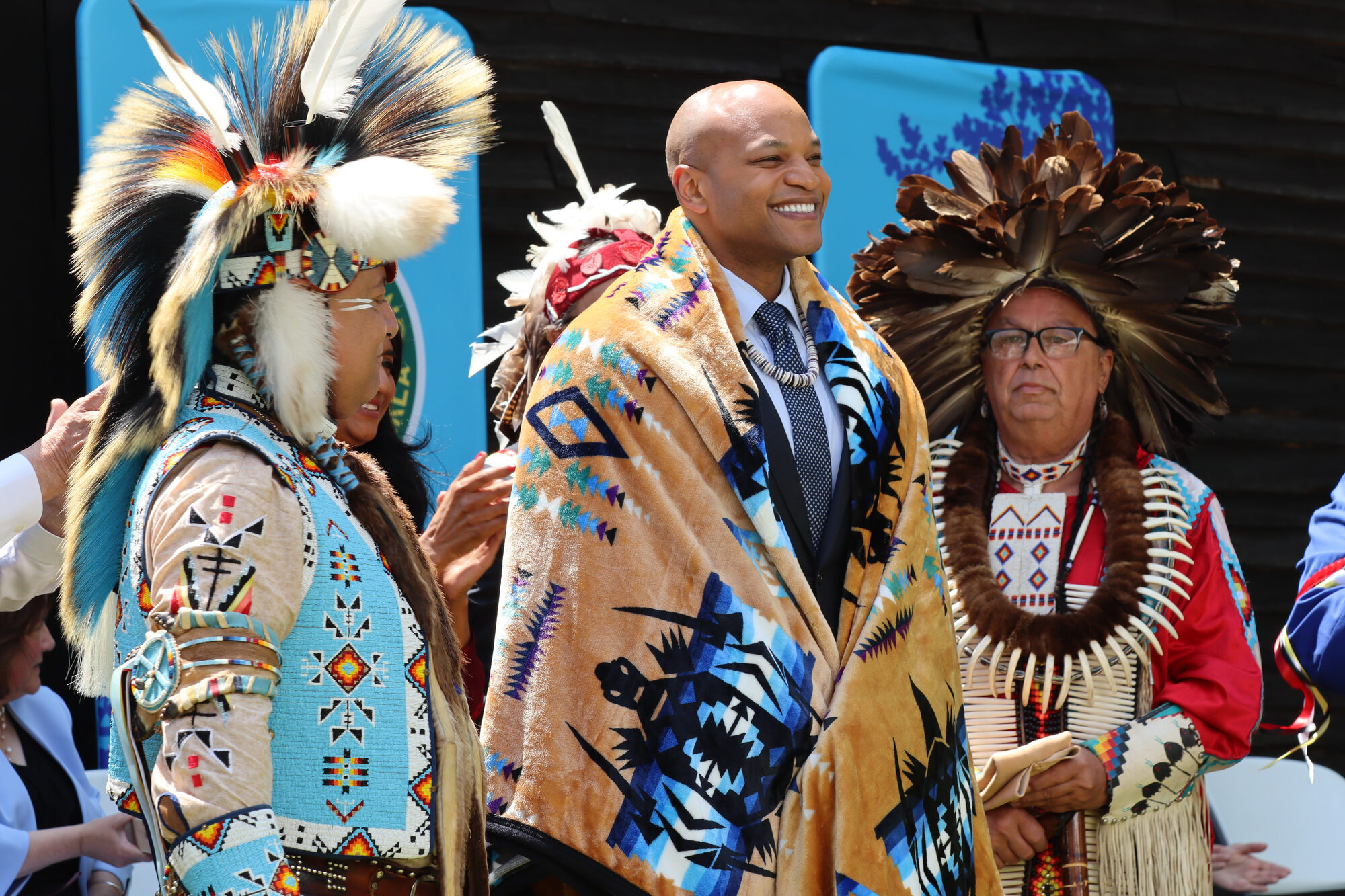 Three people in ceremonial regalia and a suit gathered at an outdoor celebration