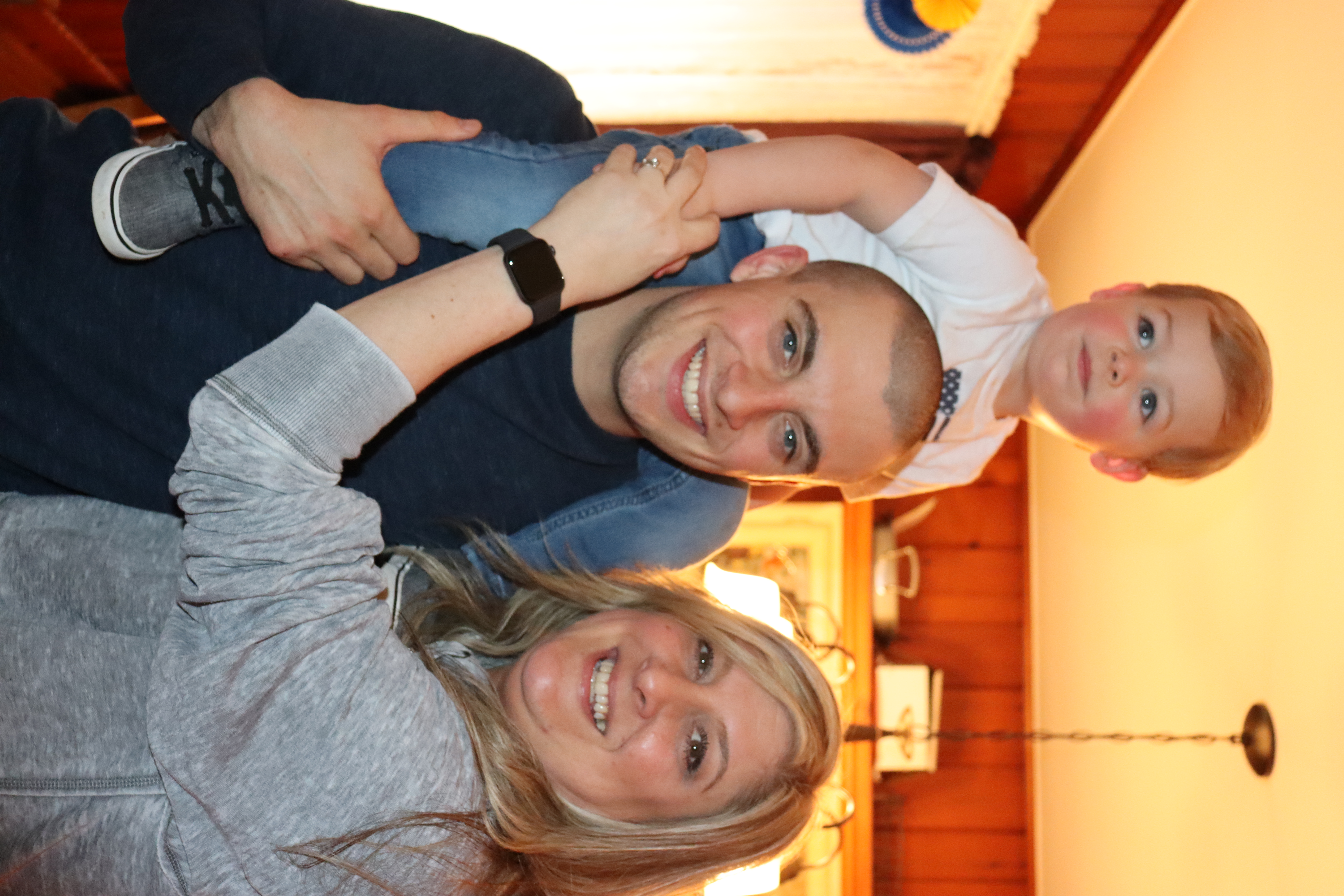 A man carries a child on his shoulders while a woman leans in beside him, all smiling