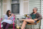 Two people sitting on a front porch, showing each other things on their phone and laptop