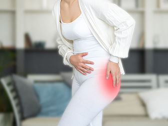 a woman slightly bent over and dressed all in white presses one hand to her stomach and the other to a pain in her left hip