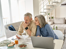 older couple with serious expressions seated at a table with food looks over papers next to a laptop