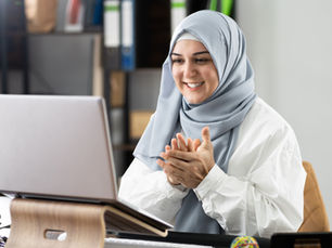 Woman wearing a light blue hijab clapping and smiling at a laptop. Office setting, neutral tones, shelves in background.