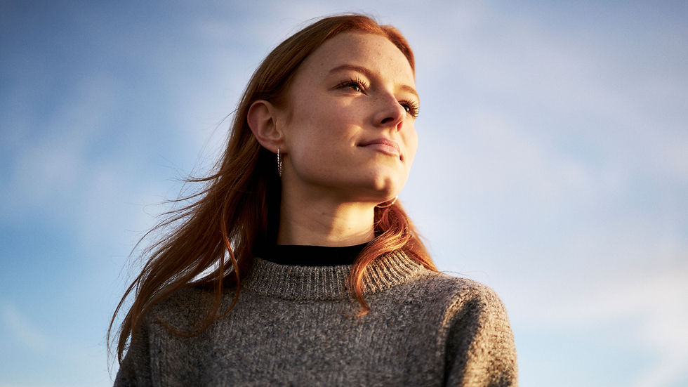 Woman with red hair gazes upward, wearing a grey sweater against a blue sky backdrop.