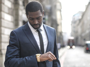 Man in a blue suit checks his gold watch on a city street with blurred buildings and traffic, exuding a focused mood.