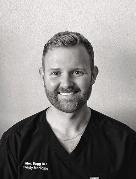 Smiling man with a beard wearing a black scrub top against a light wall.