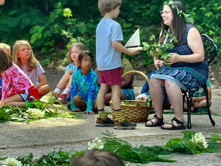 A teacher, adorned with a floral crown, shares a warm moment outdoors with her students, engaging with a child who stands before her, holding handmade items amidst a circle of attentive children.