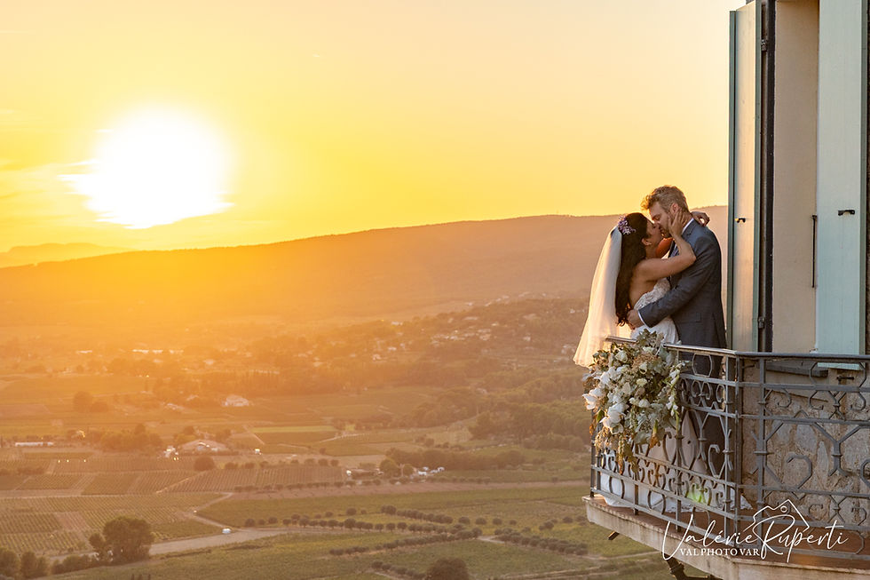 Mariés s’embrassant sur un balcon fleuri au coucher du soleil, vue panoramique sur une vallée dorée.