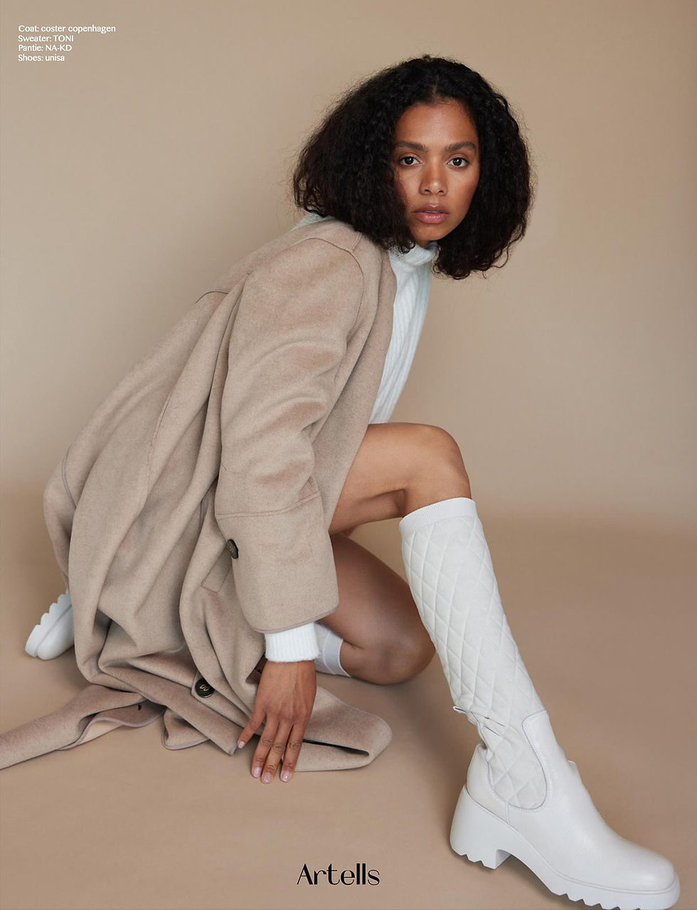 Fashion-Editorial von Andreas Liedl im Fotostudio: Model mit lockigem dunklem Haar, beigefarbenem Mantel und weißen Stiefeln vor beigem Hintergrund.