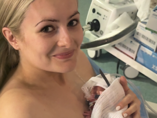 Smiling woman in a hospital bed holds a tiny newborn wrapped in a blanket, with medical equipment nearby.