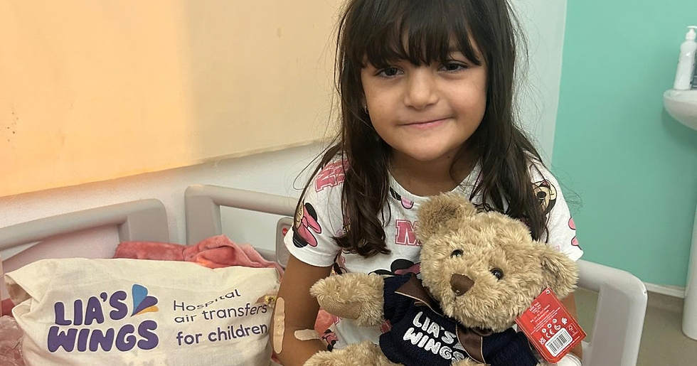 Young girl in a hospital room hugs a teddy bear wearing LIA'S WINGS shirt beside a pillow reading Hospital air transfers for children