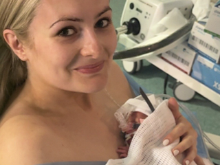 Smiling woman in a hospital holding a newborn wrapped in a white blanket. Medical equipment and supplies visible in the background.