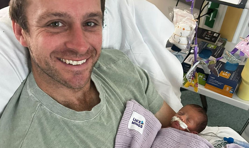Smiling man in a hospital chair cradles a sleeping newborn wrapped in a purple LIA'S WINGS blanket, with baby gifts nearby.