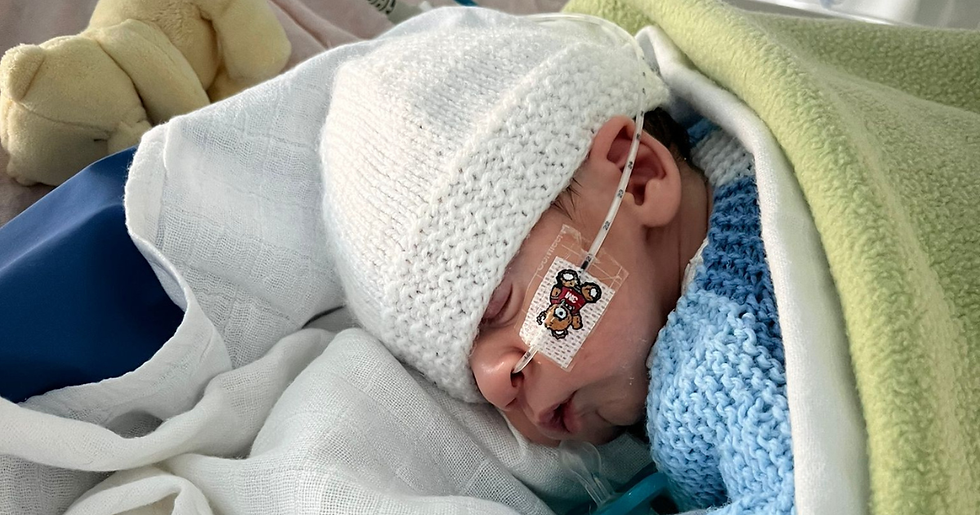 Newborn baby wearing a white knit hat, with a nasal tube taped to the face. Swaddled in blue and green blankets, sleeping peacefully.