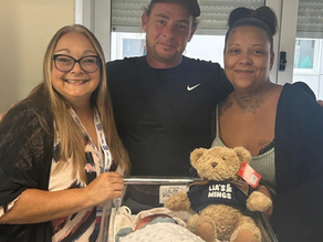 Three smiling adults stand with a newborn in a hospital bassinet beside a teddy bear shirt reading LIA'S WINGS.