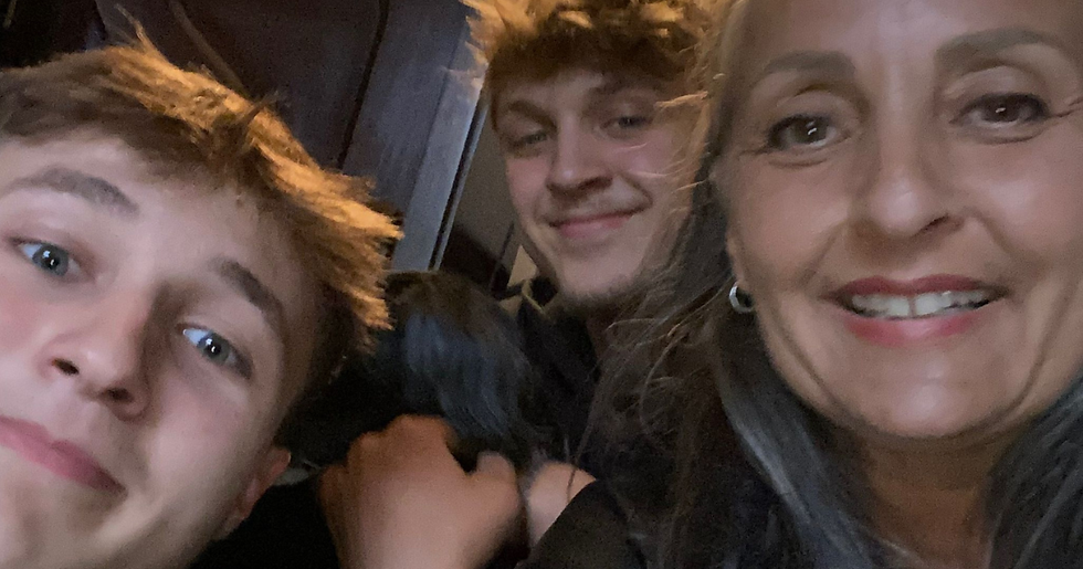 Smiling woman and two teen boys pose for a close-up selfie in a dim indoor setting, likely a car or train.
