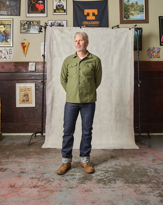 Person standing in front of a studio backdrop wearing a green overshirt, cuffed jeans, and boots