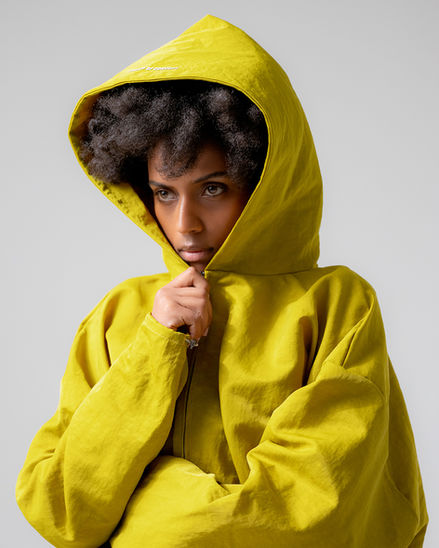 Person wearing a bright yellow hooded jacket, by Proof of Concept looking intently forward. This image, produced by Ex Litore, highlights a bold, modern fashion statement with a focus on vibrant colors and thoughtful expression.