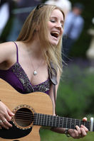 Charlie Barker playing acoustic guitar and singing live at an outdoor event, wearing a purple dress with long blonde hair. 