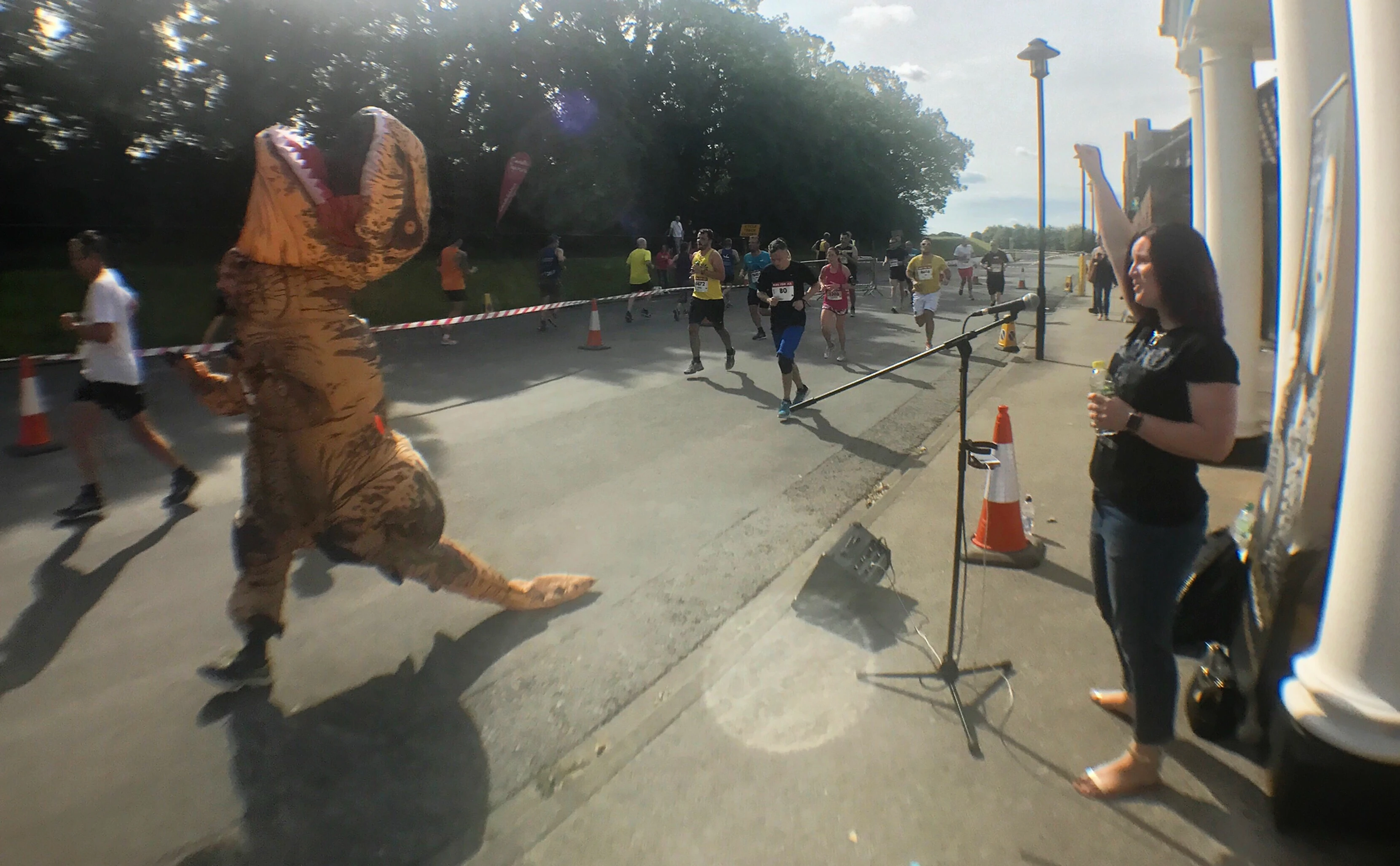 Charlie singing and cheering on the runners of York's Run For All 10K race. Running past her is a person wearing a T-Rex outfit.