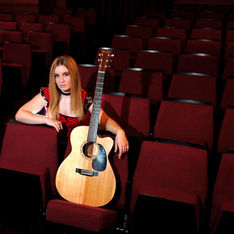 Musician Charlie Barker sat on red coloured theatre seats holding her Martin acoustic guitar, looking over to her right. Charlie is wearing a red and black top. She has long blonde hair. 