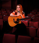 Musician Charlie Barker sat on red coloured theatre seats holding her Martin acoustic guitar, looking over to her right. Charlie is wearing a red and black top with a long black fishtail skirt. She has long blonde hair. 