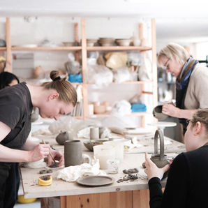 Workshop participants shaping clay pieces on wooden shelves during a ceramics class at Enaes Studio.