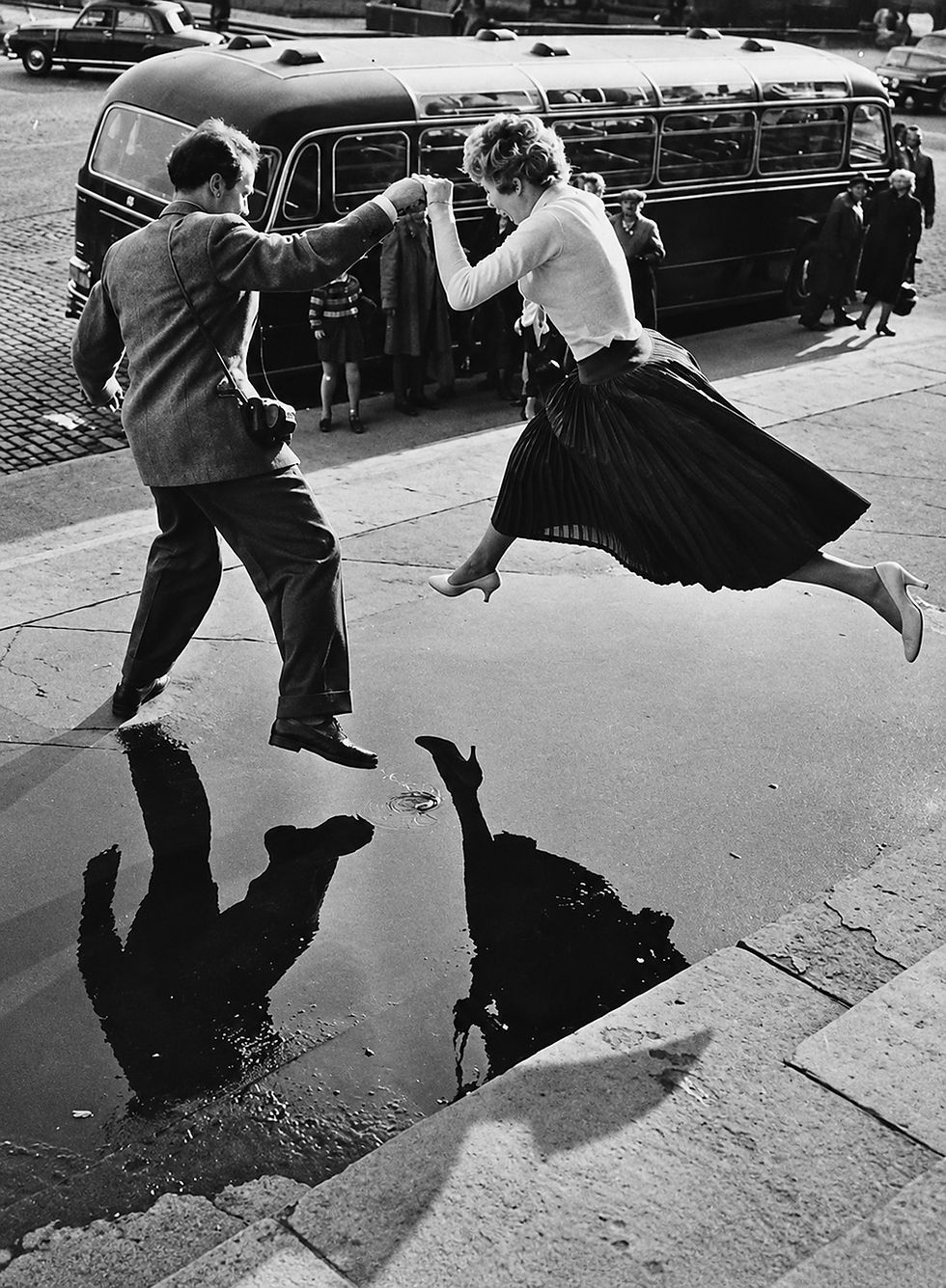 Henri Cartier-Bresson, Paris, France (Woman Jumping over a Puddle), c. 1960. A man offers his hand as a young woman, skirt flying and heels improbably airborne, leaps across a puddle on a Paris street. Below them, their dark reflections repeat the gesture, transforming an ordinary act of assistance into a moment of choreography. The bus, cars, and waiting pedestrians anchor the scene in everyday city life, while Cartier-Bresson freezes the woman at the instant when neither foot touches the ground. It is a delightful expression of his celebrated decisive moment: movement, geometry, chance, and human interaction briefly becoming one.
