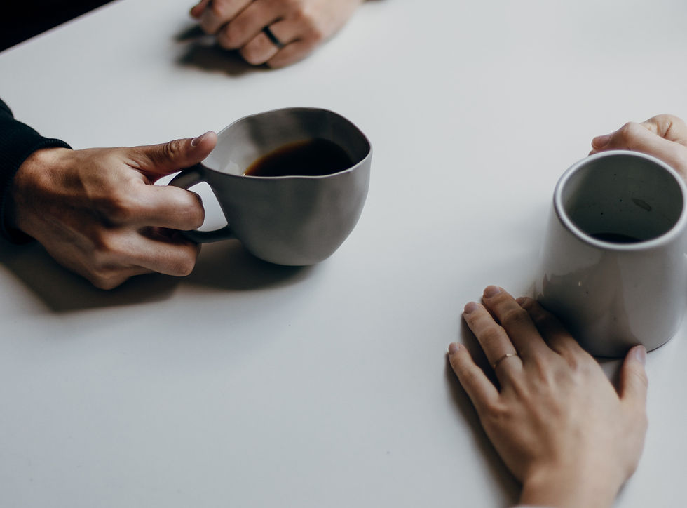 Two hands holding mugs on a white table, suggesting a relaxed conversation. The mugs are gray, and the setting is minimalistic.