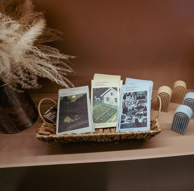 Retail shelf displaying 'A Place Here' recipe booklets in a woven basket, wooden rainbow decorative objects, and cookbooks, with dried pampas grass in a vase