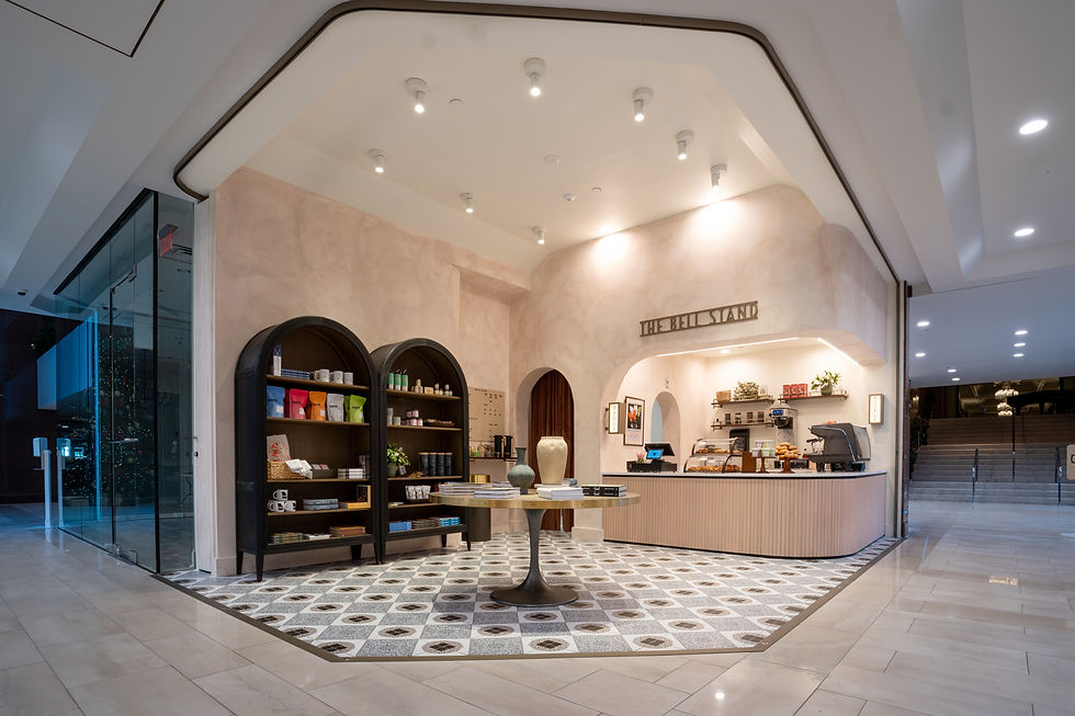 Interior of THE BELL STAND café and storefront with modern and stylish design. Features arched shelves on the left displaying tea, coffee, and other products. Center has a round table with books and decorative objects. Right side includes a counter with baked goods and a coffee machine. Intricate tile pattern on the floor adds to the aesthetic appeal.