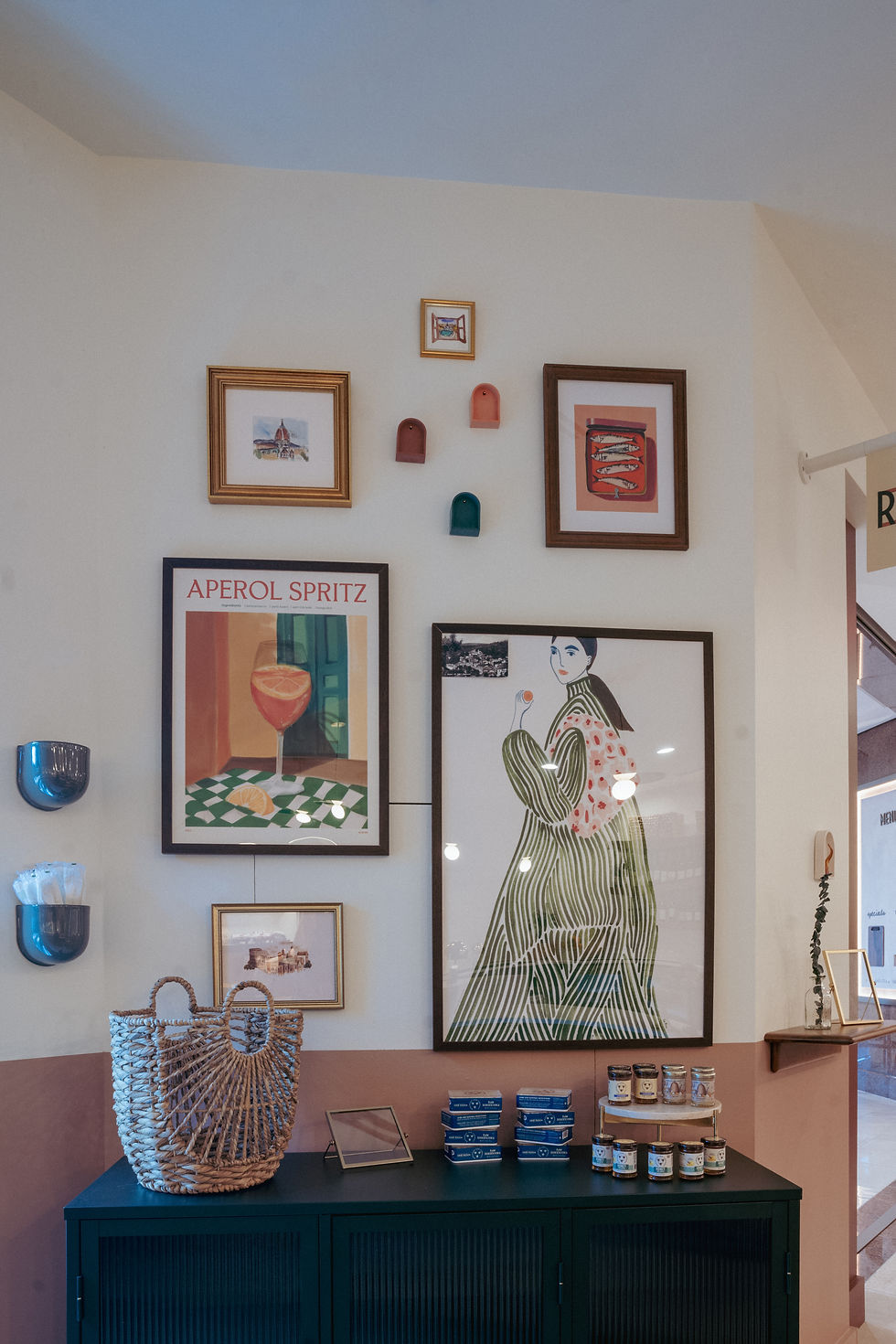 Gallery wall featuring framed artwork including an Aperol Spritz poster and art print, with small decorative wall vases and a table display of books and a woven basket below.